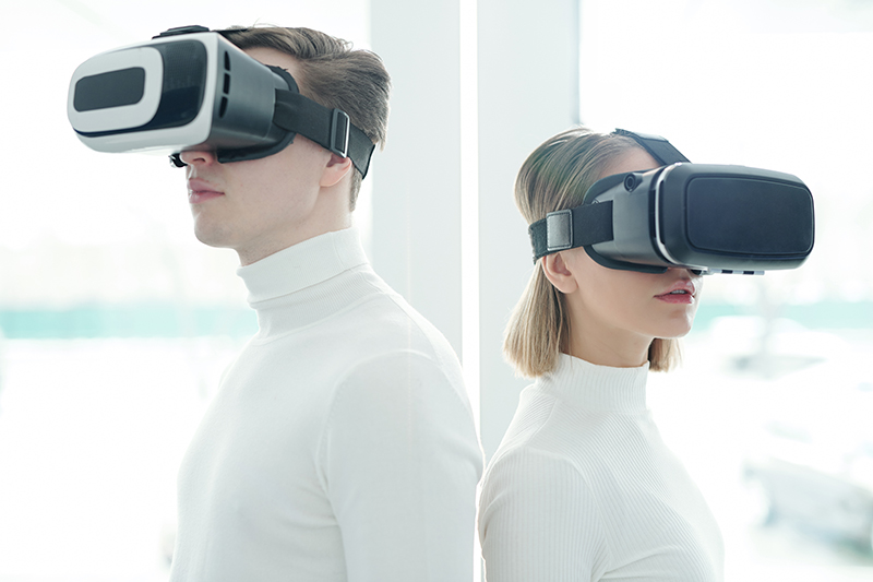 Young people in white sweaters wearing virtual reality goggles standing back to back
